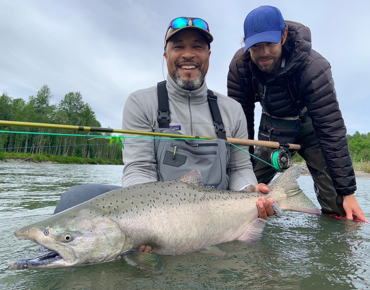 Två sportfiskare i vadare håller en stor prickig lax i en älv, båda visar stolthet över sin fångst under ett fiskeäventyr.