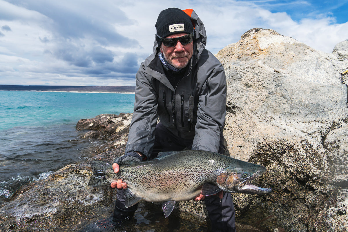 En sportfiskare i grå jacka och solglasögon står på klippig strand, håller en stor fisk framför turkost vatten och molnig himmel.