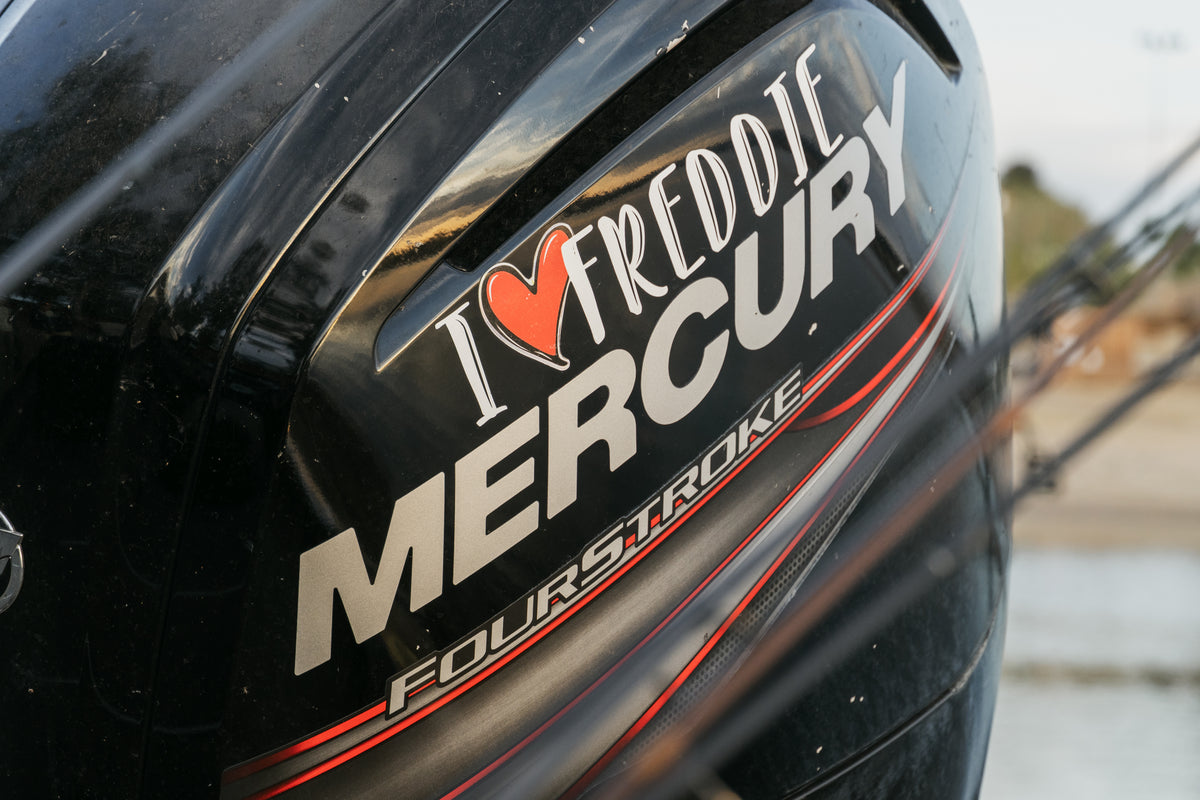 Närbild på svart Mercury FourStroke båtmotor med I ♥ FREDDIE, perfekt för sportfiskare på fiskeresa. Vatten och strand i bakgrunden.