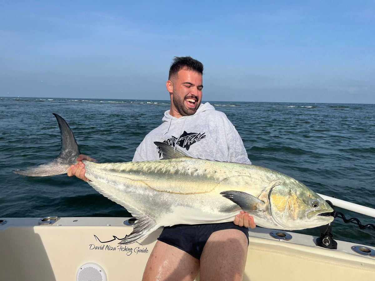 En man sitter i en båt, håller en stor fisk och ler. Hav och blå himmel syns med David Niza Fishing Guide på båtsätet.