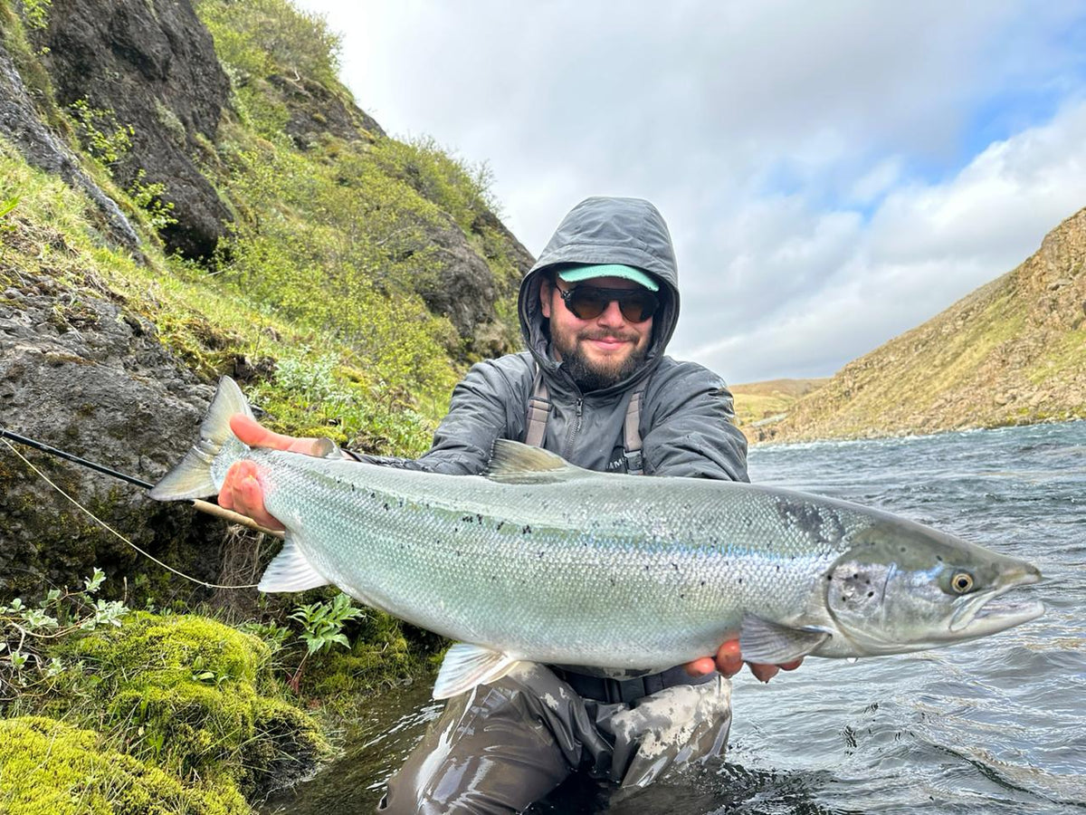 Sportfiskare med solglasögon och huva håller stor fisk vid stenig älv, omgiven av grön mossa och kullar på fiskeresa.