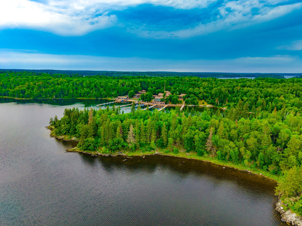 Flygfoto över en fiskecamp vid en sjö, omgiven av grön skog och klart vatten – perfekt för en inspirerande fiskeresa.