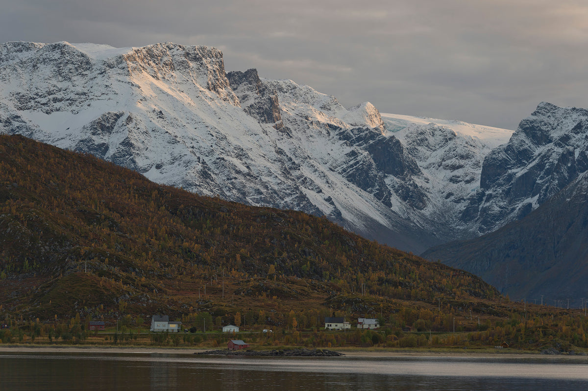 Snötäckta berg reser sig bakom skogsklädda kullar och hus. En sportfiskare njuter av fiske i lugnt vatten med vacker utsikt.