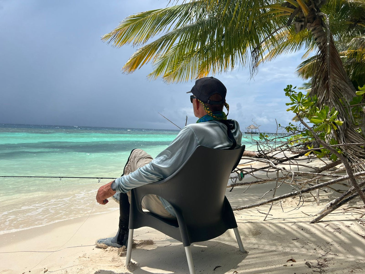 En sportfiskare sitter på en sandstrand under palmer, fiskespö i hand, blickar ut över turkost hav och njuter av lugn fiskeresa.