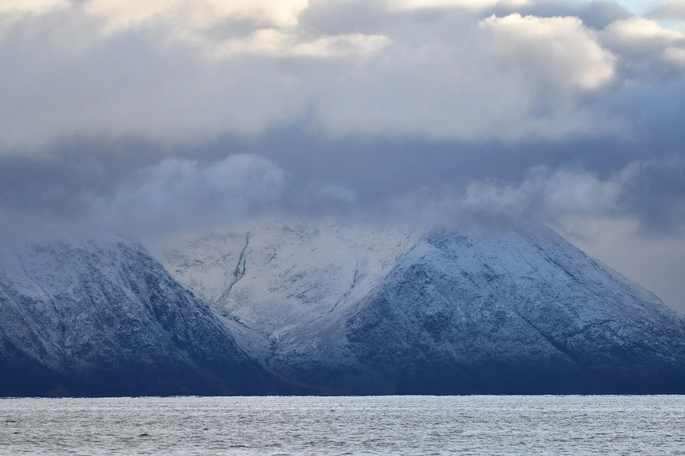 Snötäckta berg delvis dolda av moln reser sig bakom lugnt vatten under molnig himmel - perfekt för fiske och fiskeresa.