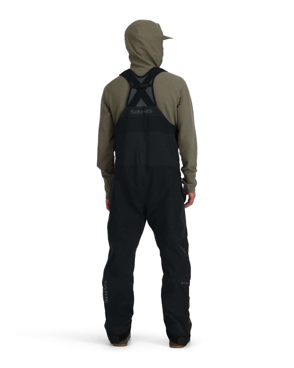 Person med ryggen mot kameran i khaki hoodie och svarta Simms GORE-TEX Pro fiskeoveraller, redo för fiskeresan.