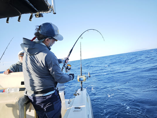 Sportfiskare i blå jacka och keps fiskar från båt på öppet vatten, drar upp fångst. Medresenär njuter av fiskeresan.