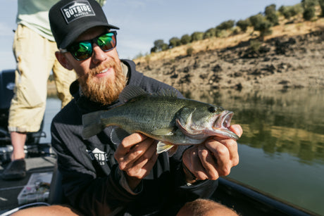 Skäggig sportfiskare med keps håller en largemouth bass i båten i Extremadura