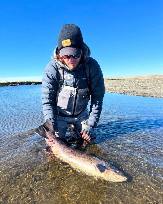 En sportfiskare i vadarbyxor och dunjacka knäböjer i vattnet och håller en stor lax - ett minnesvärt ögonblick från fiskeresan.