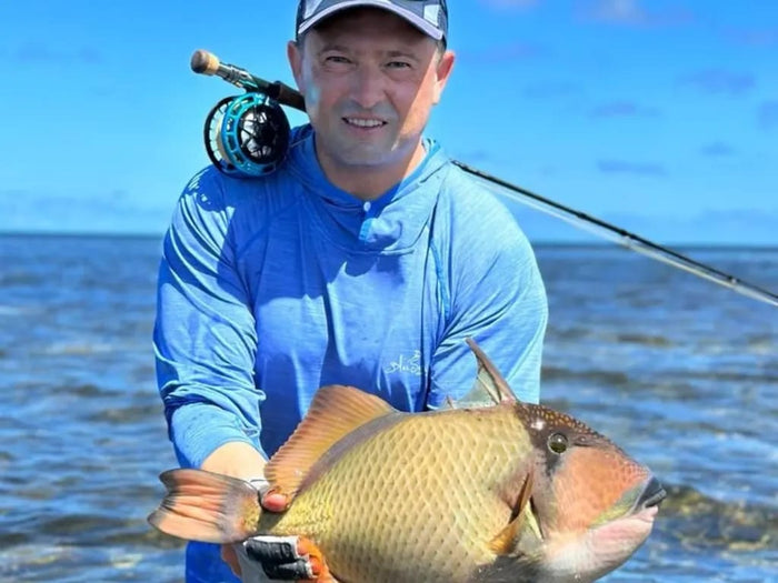 En sportfiskare i blå skjorta och keps står leende i grunt vatten med en stor triggerfish. Fiskespö vilar på axeln under klar himmel.