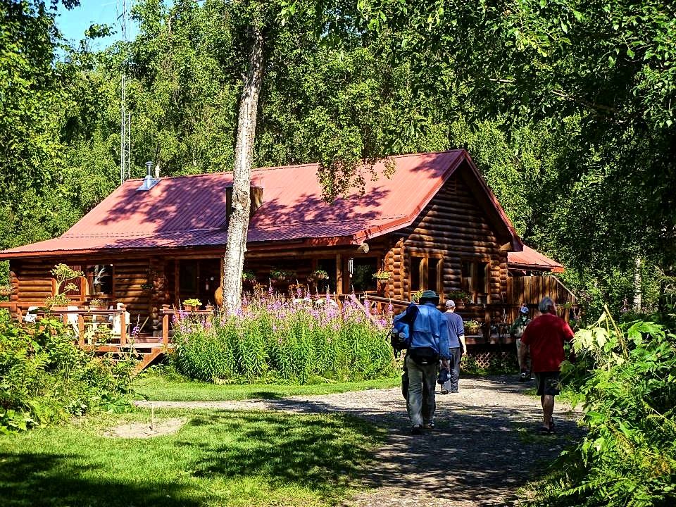 En grupp redo för sin fiskeresa går mot en timmerstuga med rött tak, omgiven av grönska och höga träd en solig dag.