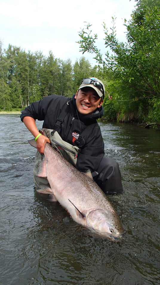 Sportfiskare i vadarbyxor står leende i en grund älv med stor fisk, omgiven av grönska - perfekt för fiskare.