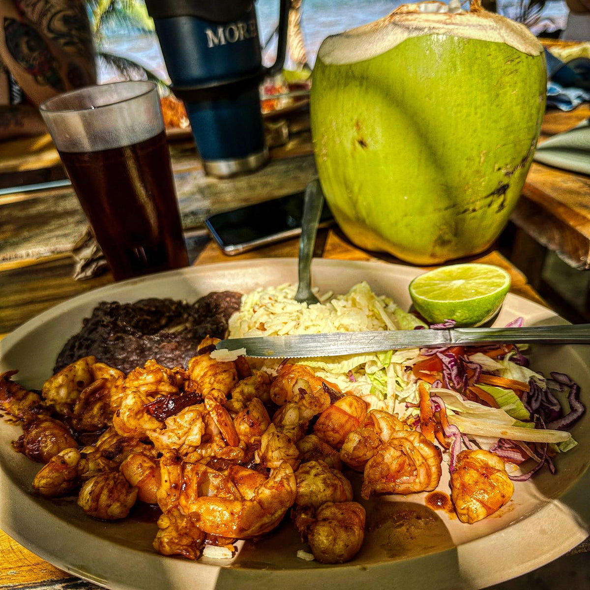 Tallrik med räkor, svarta bönor, coleslaw, sallad och lime för sportfiskare, serverat med kokosdryck och mörk dryck på soligt bord.