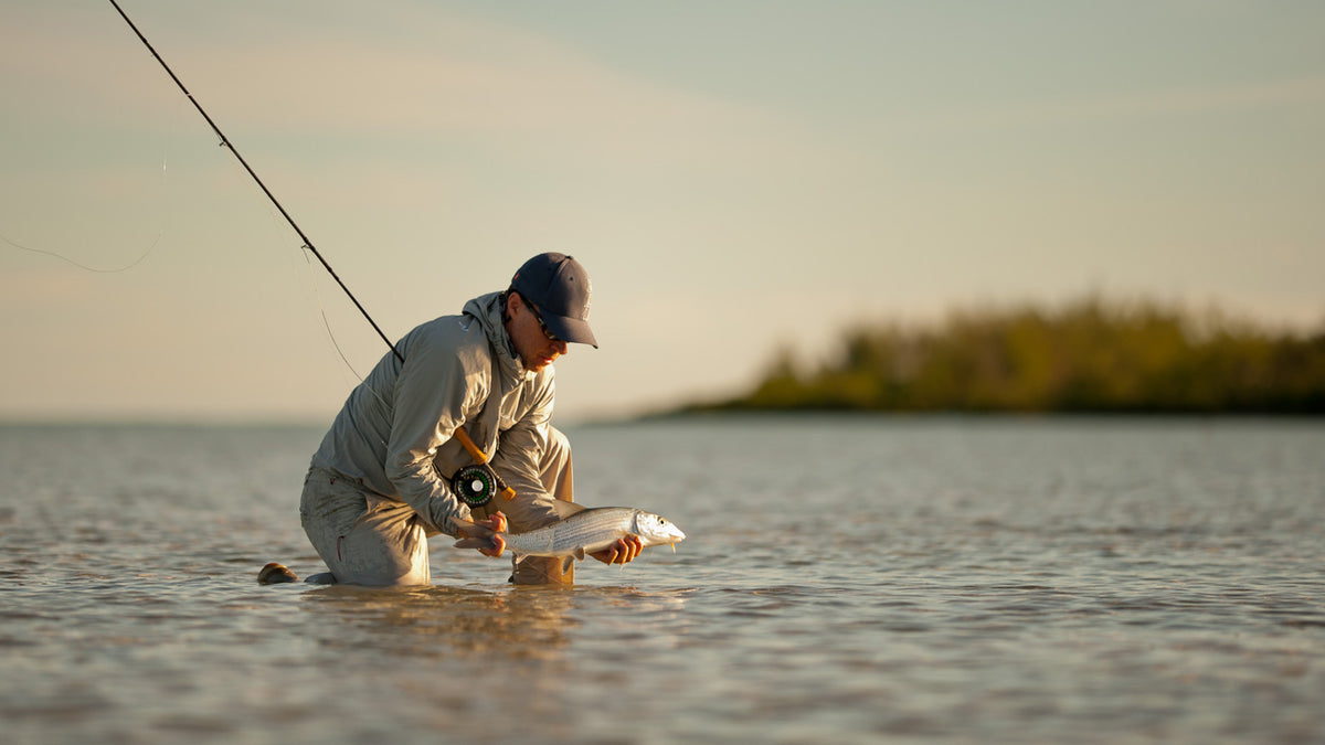 Sportfiskare i keps och fiskeutrustning knäböjer vid strand, håller fångad fisk, spö på axeln, lugn sjö och träd i bakgrunden.