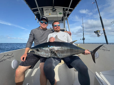 Två sportfiskare på båt håller en stor tonfisk och ler mot kameran. Spön syns mot blå himmel och lugnt hav under fiskeresan.
