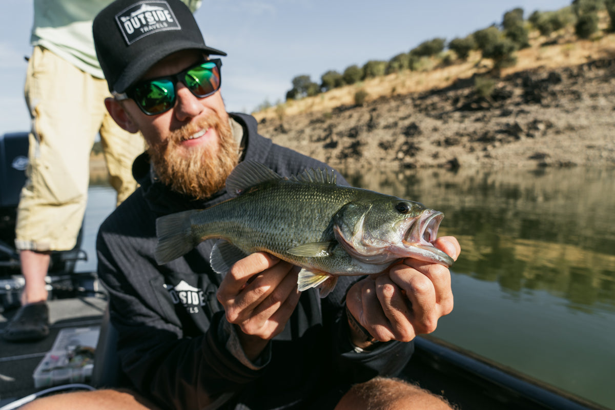 Skäggig sportfiskare med solglasögon och svart keps håller en largemouth bass i båt på lugn flod med klippstrand och träd.