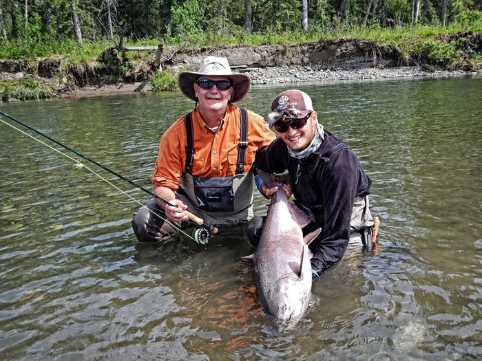 Två sportfiskare i vadande fiskeutrustning håller en stor fångad fisk, med träd och grön flodbank-idealisk fiskeresa.