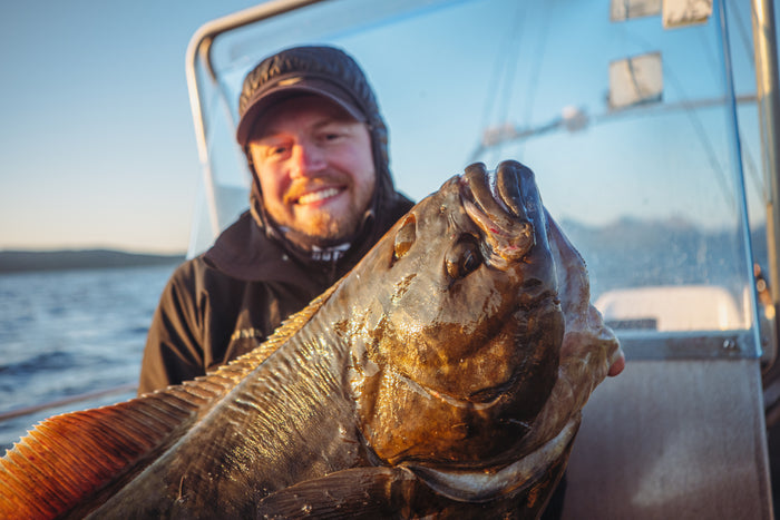 En leende sportfiskare i outdoor-kläder håller en stor fisk på en båt med vatten och avlägsen kustlinje i bakgrunden.