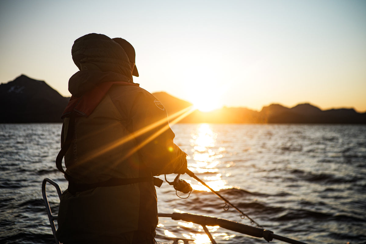 Sportfiskare i jacka står på en båt i gryning eller skymning med fiskespö, solreflektion på vattnet och siluett av berg.