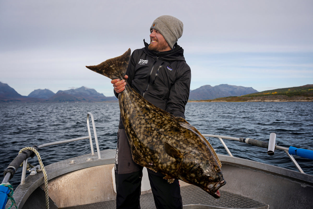 Sportfiskare med grå mössa och svart jacka visar stolt upp stor platt fisk på båt, berg och vatten i bakgrunden.