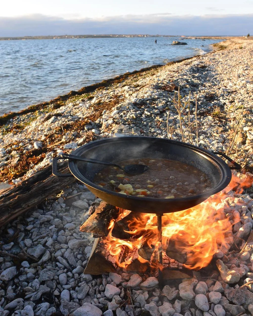 Svart gryta med gryta kokar över öppen eld på klippstrand, hav och kust syns i bakgrunden-idealisk efter fiskeresa.