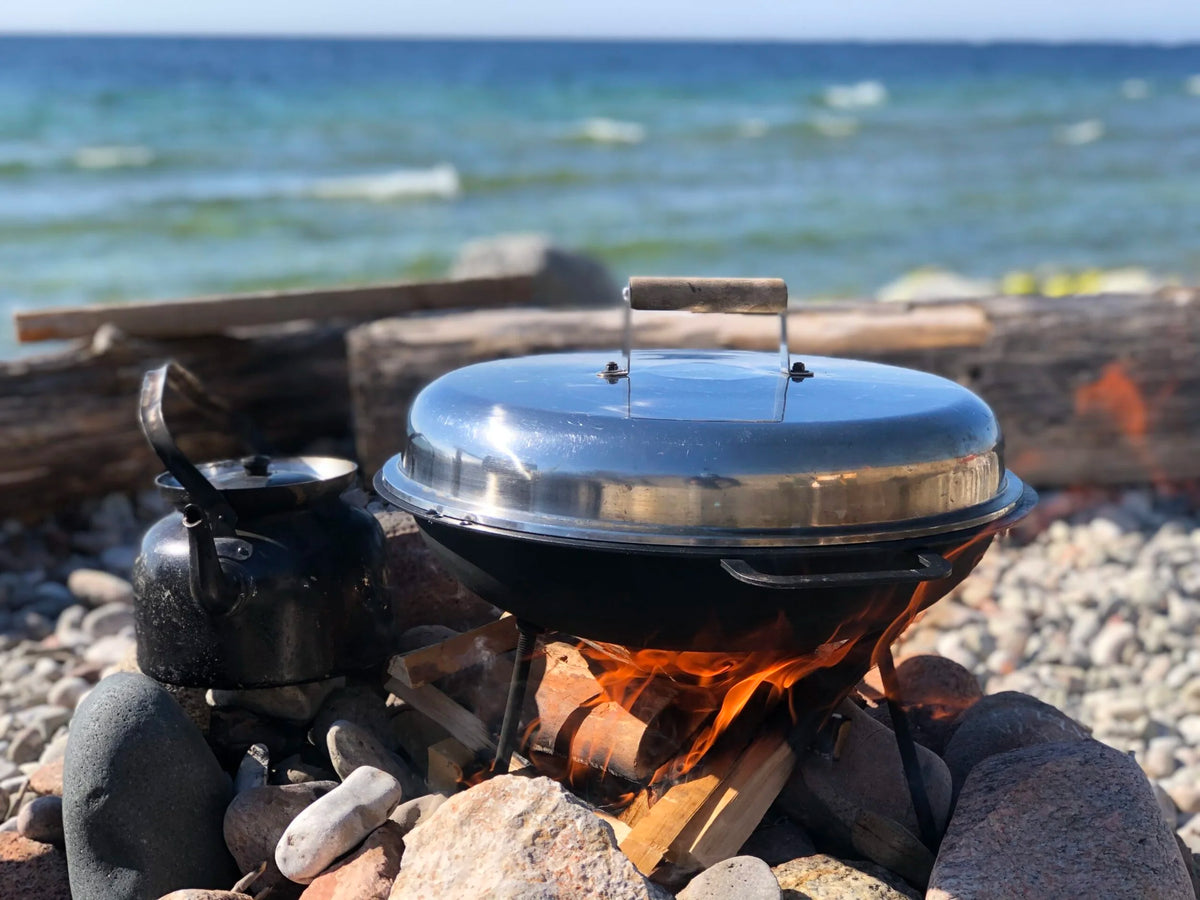 Metallkittel och gryta över lägereld på stenstrand med drivved och vågor - inspiration till avslappnande fiskeresa vid havet.