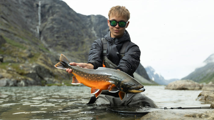 En sportfiskare med solglasögon och vadarbyxor håller en stor färgglad fisk vid ett klippigt fjällvatten under fiskeresan.