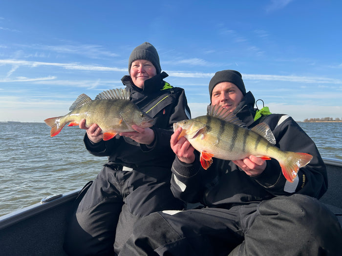 Två sportfiskare i varma kläder håller upp nyfångad fisk med orange fenor i båt, leende mot kameran på lugnt vatten.