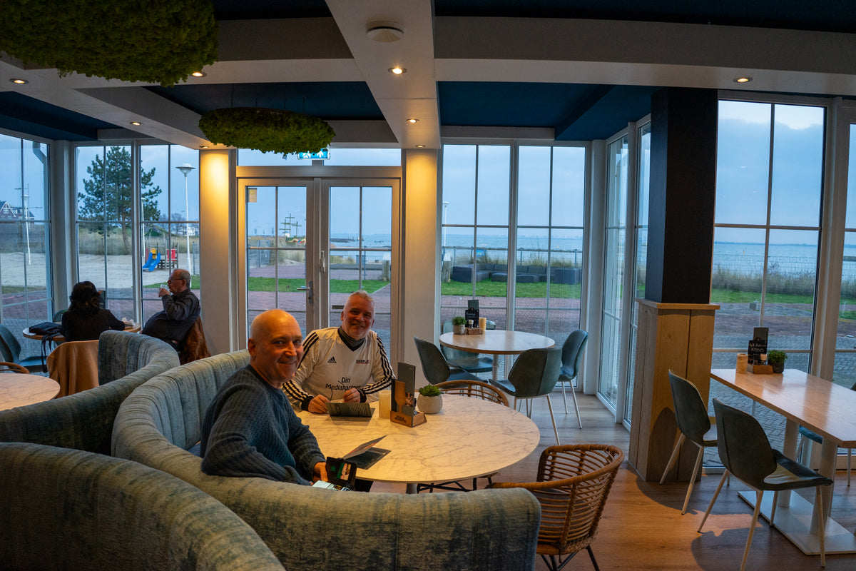Två män samtalar leende om sin senaste fiskeresa vid ett runt bord i café med havsutsikt, strand och andra gäster.