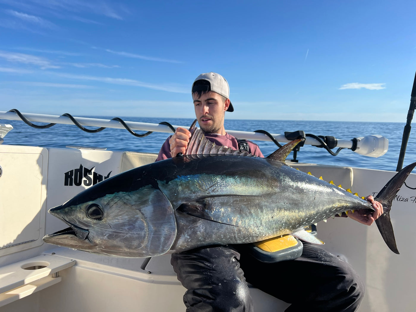 Sportfiskare med vit keps och hängselbyxor poserar på båt med stor tonfisk i knät, havet och blå himmel i bakgrunden.
