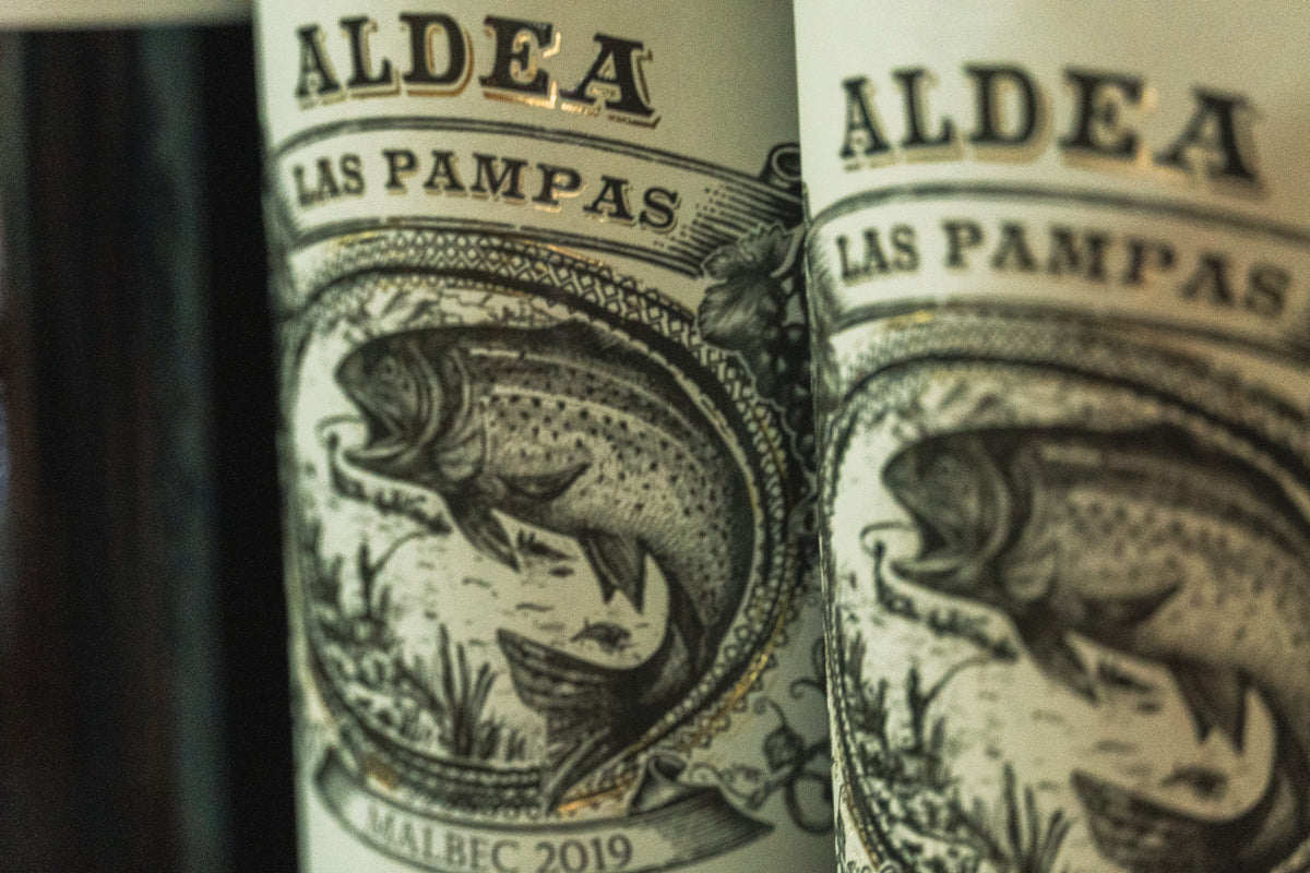 Närbild av vinflaskor med ALDEA LAS PAMPAS-etikett, fisk- och landskapsmotiv, Malbec 2019, inspirerat av fiskeresa.