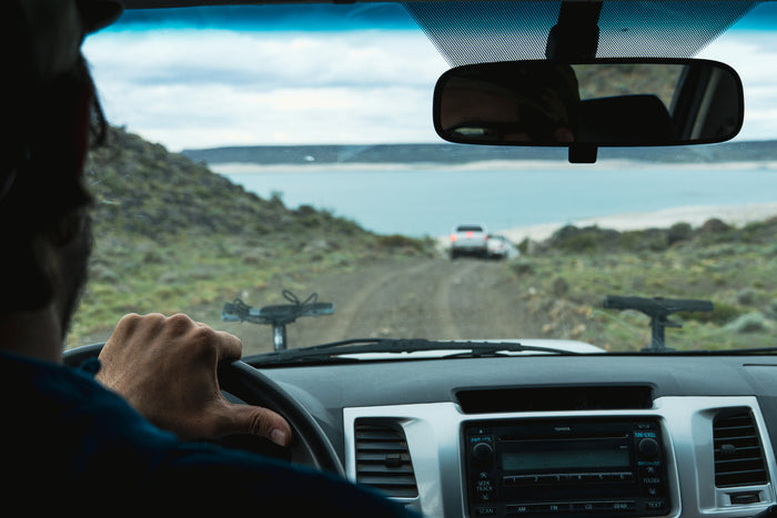 Vy från bilen visar sportfiskares hand på ratten mot sjön, grusväg, vit bil framför, fiskeresa bland klippor och moln.