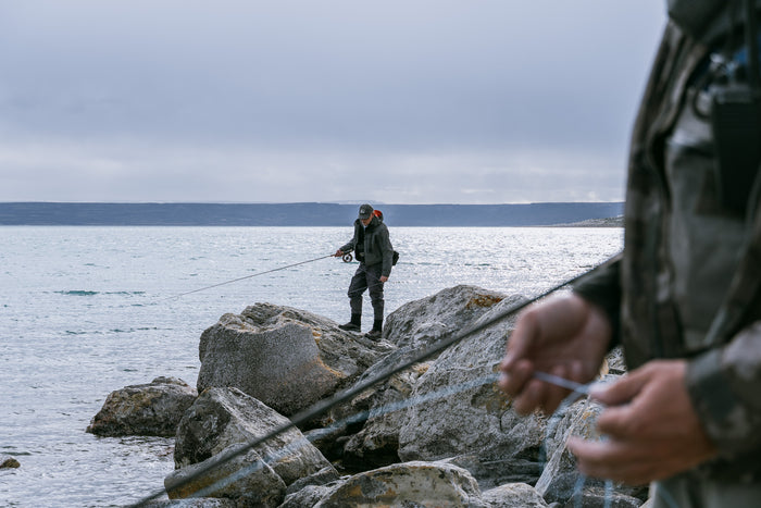 Två sportfiskare flugfiskar vid en klippig kust; en kastar ut från stenarna, den andra syns suddigt i förgrunden med fiskeutrustning.