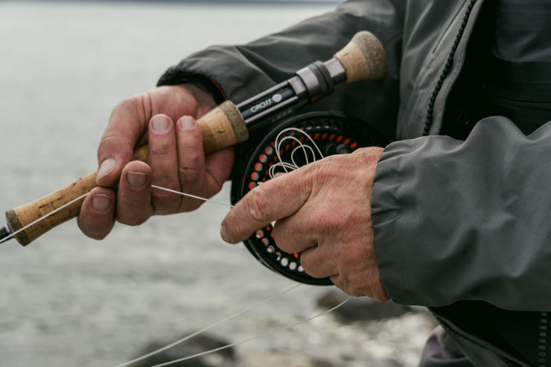 Närbild av sportfiskares händer som trär linan på spö och rulle. Grå jacka, vatten i bakgrunden under fiskeresan.