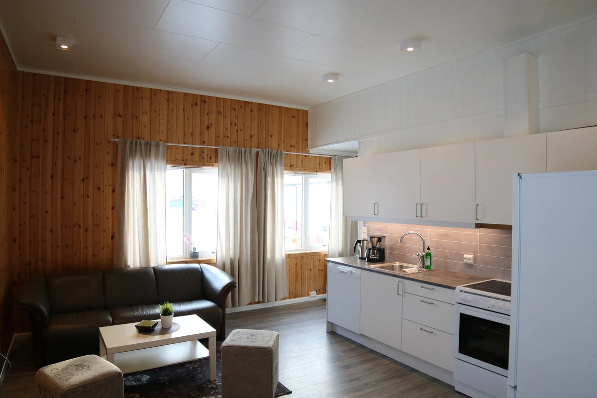 Trivsam lounge med träpanel, mörk soffa och två ottomaner för sportfiskare. Intill finns modernt vit kök, stora fönster.