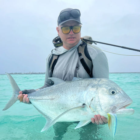 Sportfiskare på fiskeresa i Maldiverna håller stor silverfisk med betesfisk, står i turkost vatten med spö på ryggen.