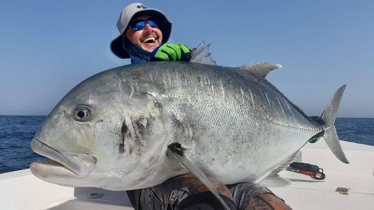 En glad sportfiskare med solglasögon, hatt och handskar håller en stor fisk ombord på en båt under en minnesvärd fiskeresa.