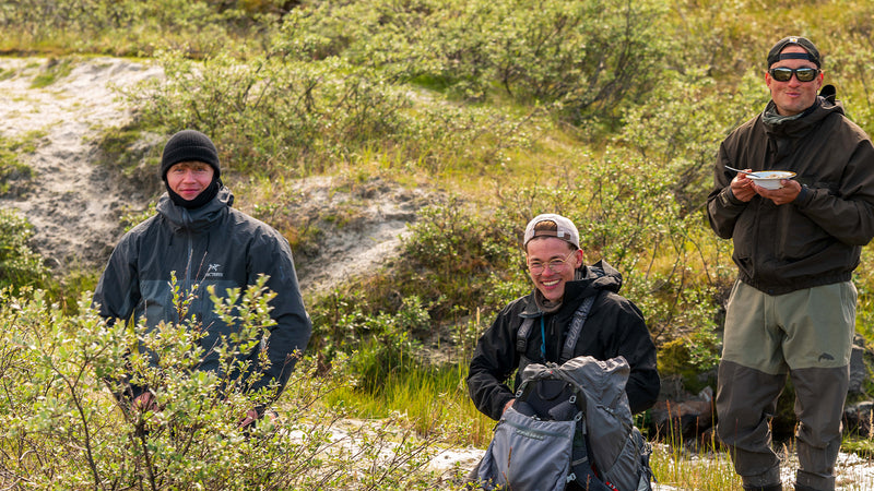 Tre personer i friluftskläder vid gröna buskar, en med skål och sked, ler mot kameran under en solig fiskeresa i naturen.