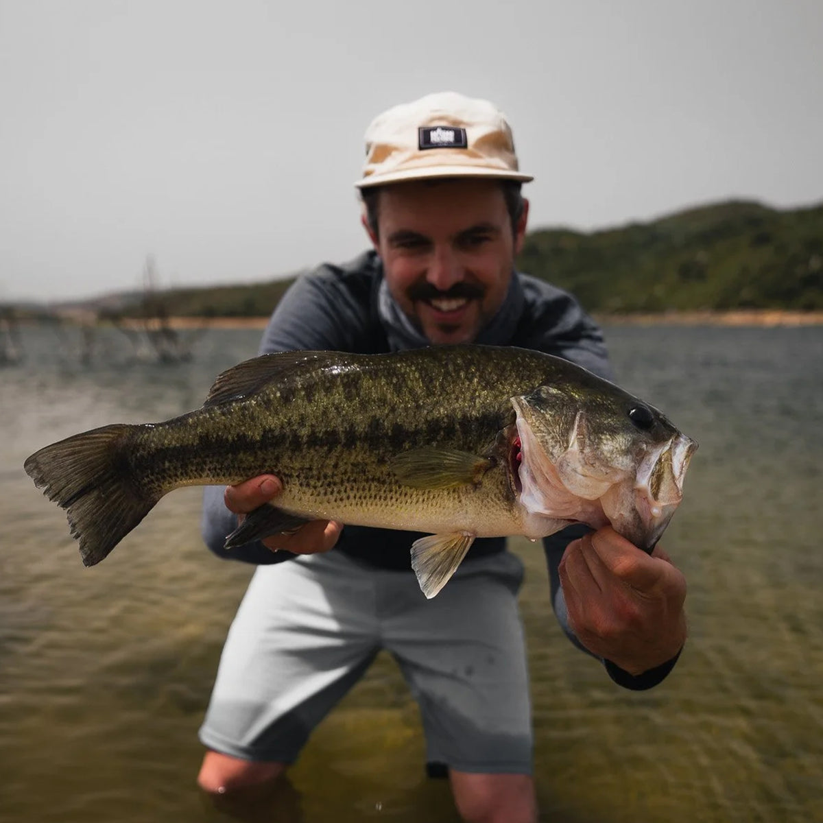 Man i hatt vid sjö håller stor fisk, glad under fiskeresa Sardinien Italien. Gröna kullar och molnig himmel.