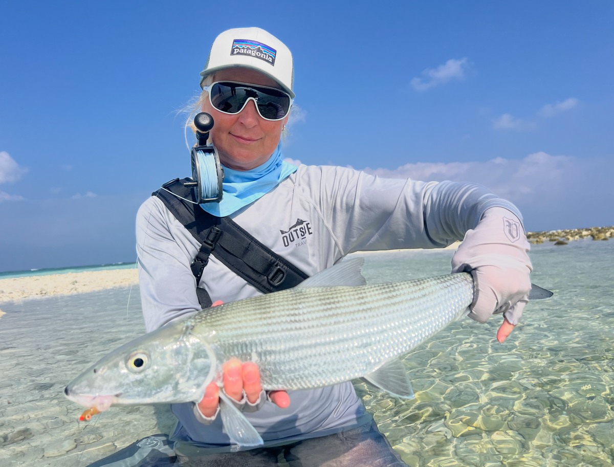 Sportfiskare med solglasögon, hatt och fiskeutrustning håller en stor silverfisk i klart grunt vatten vid sandstrand under blå himmel.