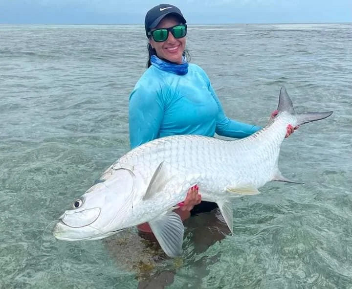 En leende sportfiskare med solglasögon, blå skjorta och keps står i grunt vatten och håller en stor silverfisk med båda händer.
