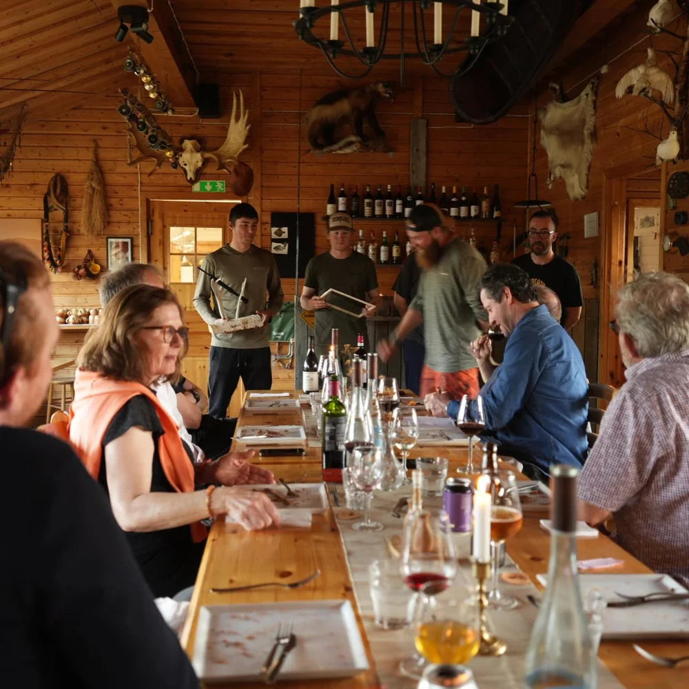 Sportfiskare samlas runt ett långbord för middag i en träpanelad stuga med djurhudar, flaskor och serverande gäster.
