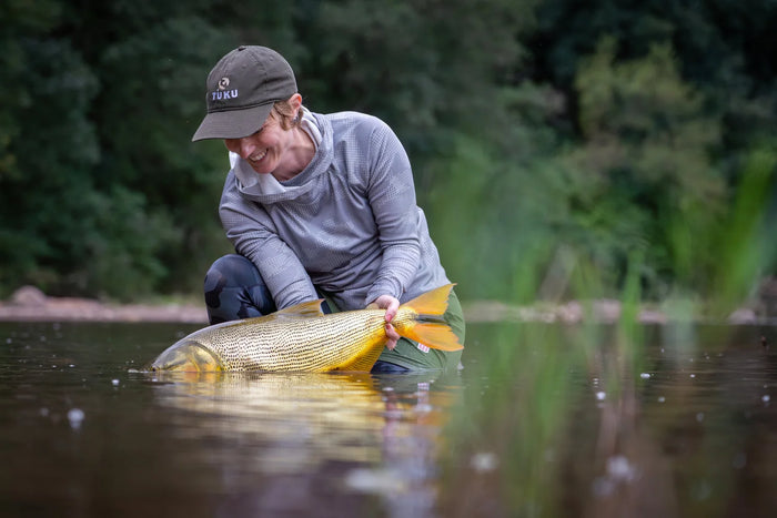 En person knäböjer i grunt vatten och ler med en stor gyllene fisk under en fiskeresa; suddig skog i bakgrunden.