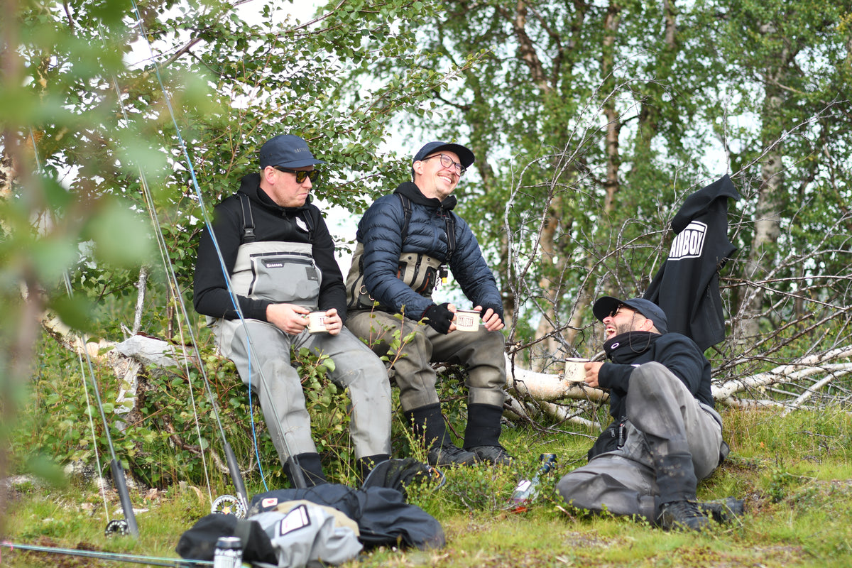 Tre sportfiskare i fiskeutrustning sitter på gräs och stockar, pratar med muggar i handen under fiskeresa bland skog och utrustning.