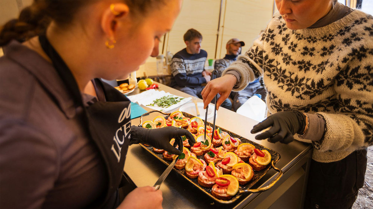Två personer förbereder matbrickor med grönsakspajer i en matbod. Tre sportfiskare i tröjor följer scenen i tältet.