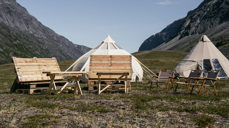 Två canvastält på gräs mellan fjäll, med bänkar, bord och campingstolar-idealisk plats för sportfiskare efter fiskeresa.