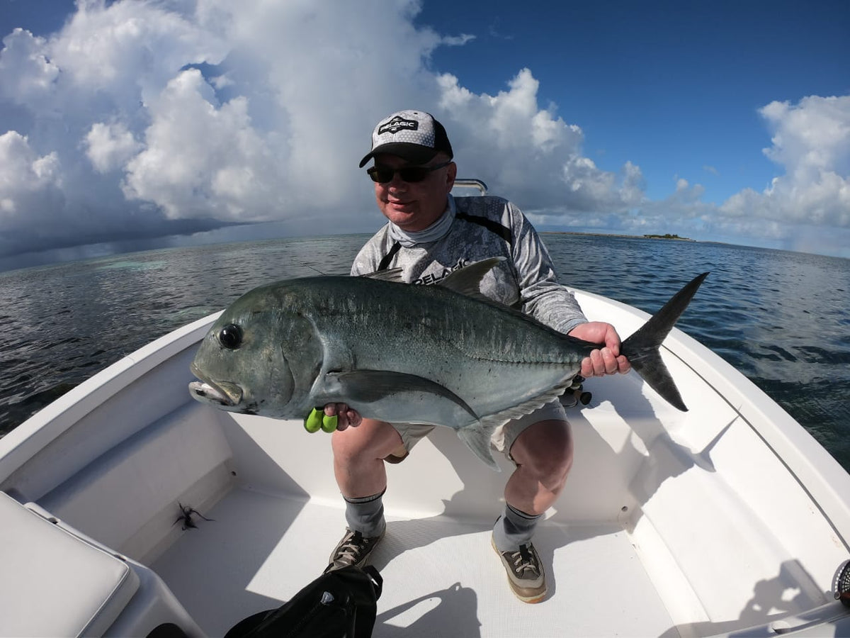 Sportfiskare med hatt och solglasögon i båt håller stolt en stor blank fisk. Lugnt vatten speglar ö med molnig himmel.