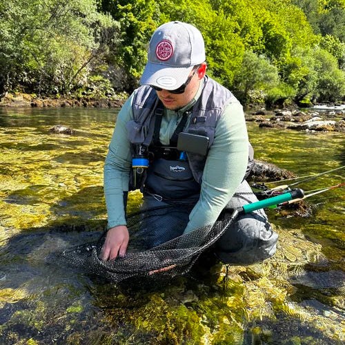 Sportfiskare i fiskeutrustning knäböjer vid klar grund älv, håller håv, släpper ut fångst bland gröna träd på spännande fiskeresa.