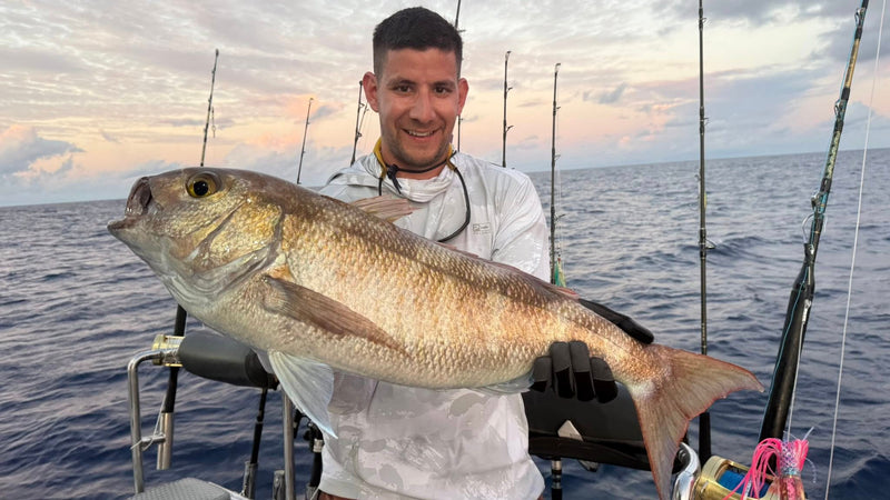 En sportfiskare i vit skjorta och handskar håller en stor fisk på en båt, fiskespön syns och havet breder ut sig i solnedgången.